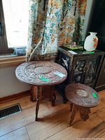 Both carved wood side tables shown together near stained glass cabinet and window with decorative curtain.