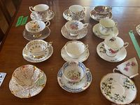 Top view of all 12 tea cups and saucers arranged on wooden table, showing varied floral designs and gold trims.