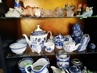 Display of the blue and white porcelain set on a shelf, showing teapot, bowls, small containers, and decorative pieces including a shoe and a cat figurine.