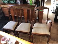 Set of three wooden dining chairs including one armchair, showing carved backs and light striped fabric seats