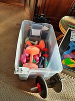 Clear plastic bin containing several colorful plastic dumbbells and two metal dumbbell bars with iron weight plates set on the carpeted floor.