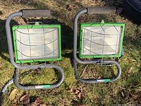 Pair of green framed Haussmann work lights with protective wire guards outdoors on grass with some leaves, showing front and stands.
