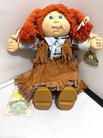 Front view of 1983 Cabbage Patch Cowgirl Doll showing orange yarn hair in pigtails, soft vinyl face, brown fringed cowgirl outfit, and brown shoes. Tags attached including original Cabbage Patch Kids tag.