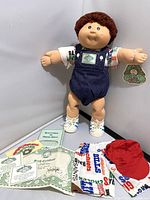 Image of 1983 Original Cabbage Patch Kid doll standing with original Pamper, sports-themed shirt and shorts, white sneakers, red cap, and accompanying documents including birth certificate and envelope.