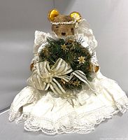 Front view of a brown plush bear dressed as an angel in an off-white lace dress holding a green wreath with gold ribbon decorations.