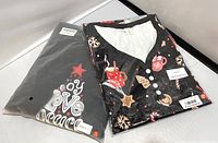 Photo of two holiday pajama sets: one folded black night dress with holiday cocoa-themed prints and buttons, one folded two-piece pajama set with plaid pants and joy-themed top in packaging.