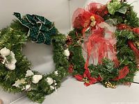 Photo showing two large decorative Christmas wreaths, one with white floral decorations and green bow, the other with red mesh and angel figurine.