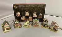 View of all six miniature building ornaments arranged in front of original box showing images and Liberty Falls branding. Ornaments have hanging cords and depict small town buildings in painted detail.