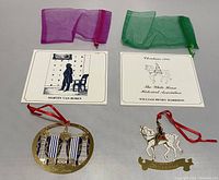 Two White House Christmas ornaments with their respective brochures and two organza bags on a plain surface. Left ornament oval shaped with blue and white curtains and chandelier; right ornament horse shaped with red hanging ribbon.