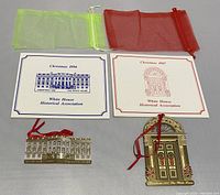 Two White House Christmas ornaments from 1986 and 1987 with matching brochures and colored organza bags on a neutral background