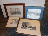 Three framed artworks shown leaning against a wall on a wooden floor and carpet. The artworks include two hand colored prints and one pencil drawing with a blue mat.