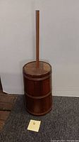 Full view of the wooden butter churn with its plunger, standing upright on carpet beside a wall.