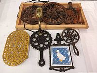 Full set of seven cast iron trivets displayed on white background with wooden tray holding three trivets at top. One trivet converted to electronic wall lamp.