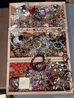 Top view of three wooden drawers filled with assorted costume jewelry mainly rings, bracelets, earrings, and necklaces, vibrant colors including orange, black, purple beads and metallic metal chains.