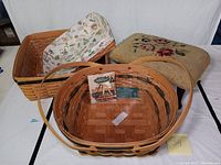 Three Longaberger woven baskets with one large basket in foreground holding a pamphlet and smaller baskets behind, alongside a vintage needlepoint foot stool with floral pattern.