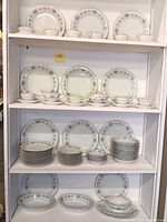 Shelves filled with multiple stacks of plates, bowls, and cups with the Noritake Allston green floral and gold pattern.