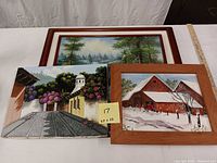 Three scenic oil paintings displayed on a table; two are framed showcasing a forest waterfall and snowy barn, the unframed one shows a cobblestone street with flowers and a white church tower.