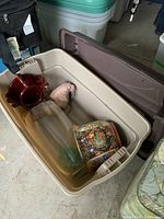 Open plastic storage bin showing multiple decorative items inside including a red ceramic bowl, a plaster walrus, a floral jardiniere with wooden stand, and a large glass vase.