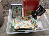 Two ceramic casserole dishes with floral designs, books including 'The Doctors Book of Home Remedies', a wine aerator, a lint brush, and a photo album arranged inside a clear plastic bin.