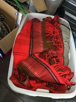 Red woven blanket with red, brown, and black stripes and fringed tassels, folded in white plastic storage tote.
