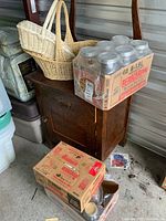Three flats of canning jars on top of and near a dark wooden cabinet, with two wicker baskets with handles placed behind them. Some jars are stored in original Bernardin cardboard boxes.