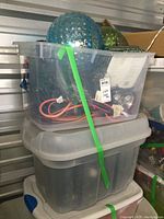 Tote filled with assorted Christmas lights and power cords with two large textured glass balls visible inside, secured with a green strap.