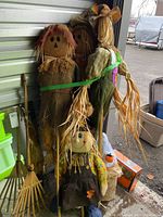 Overall view of the Halloween decorations including five straw figures, a white ghost, and some small path markers, plus two rakes leaning against the wall.