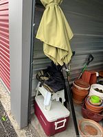 Green outdoor table umbrella folded and tied near metal wall with white two-step stool and red and white Coleman cooler below it, black carrying bag on the stool.
