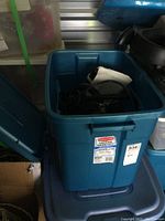 Top view of open large blue Rubbermaid storage bin with black electrical cord and attachments inside, showing steam cleaner parts.