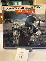 Front cover of John Cougar Mellencamp LP album 'Scarecrow' showing artist leaning on fence with title text.