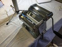 Glossy stainless steel Marcato Atlas 150 pasta maker with hand crank attached, viewed from front and side showing table clamp