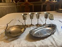 Photo showing four silver goblets, silver candy dish with handle, and ornate oval silver bread plate on table.