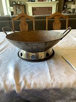 Steel wok on metal heating ring shown on dining table with wooden chairs in background.