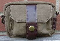 Front view of khaki camera pouch bag with leather-style strap and brass snap button, showing fabric texture and a Roots brand tag.