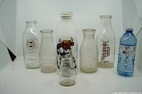 Front view of 7 vintage glass milk bottles showing various labels, embossing, and cow illustration on ceramic bottle. Plastic water bottle for size comparison.
