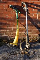 Photo showing two electric weed eaters/weed whackers standing upright against a brick wall. One unit is yellow and green with a cord wrapped around the handle, the other is silver and black without a cord and a visible protective guard near the cutting head.
