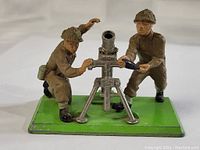 Side angle showing two painted metal soldier figures operating a silver mortar on a green base with minor wear.