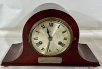 Front view of antique wooden mantle clock with Roman numeral dial and silver plaque at base.