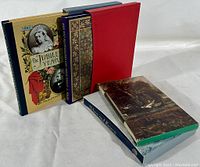 Four hardcover Folio Society books with original slip-covers arranged standing and leaning. Titles and artistic binding visible.