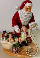 Front view showing Santa Claus holding a teddy bear and scroll with toys and gifts around base.