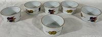 Front view of six white ceramic ramekins arranged in a line, each decorated with different hand-painted fruit patterns including blackberries, grapes, and olives with gold trim.