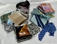 Overview of assorted scarves of various colors, patterns, and fabrics laid out showing multiple folded pieces including black floral, beige knit, blue with stripes, and purple checkered.