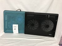Two laptop cooling pads side by side, one black with visible dual fans and buttons, the other blue with two fans under a mesh surface, both with USB connectors visible.