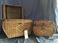 Open and closed views of a larger curved wicker chest with metal latch and decorative scroll handles and feet beside a smaller rectangular wicker chest with a latch.