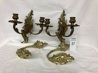 Four brass-colored wall sconces and candle holders displayed against white background. Two double-arm sconces with intricate leaf and scroll design, and two single-arm ornate holders.