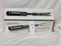 Image shows the cook's fork with built-in thermometer fully visible alongside its original box which indicates the product features and branding. The fork has long stainless steel prongs and a black digital handle with display and buttons.
