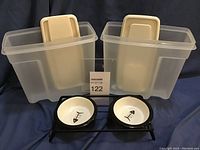 Two translucent Plastic Rubbermaid containers with hinged lids and a black metal stand holding two white ceramic pet bowls with a fish bone design.