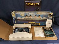 Photo showing boxed musical coin bank modeled as Titanic, paper items including a book and receipt, and a collector plate partially visible in open box.