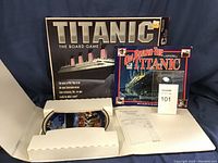 Titanic board game box and 'On Board the Titanic' story book with ticket card labeled 101 and partially visible blue collector's plate in white protective sleeve