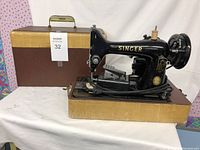 Photo showing vintage black Singer sewing machine mounted on wooden base along with accompanying brown and beige carrying case with metal handle.
